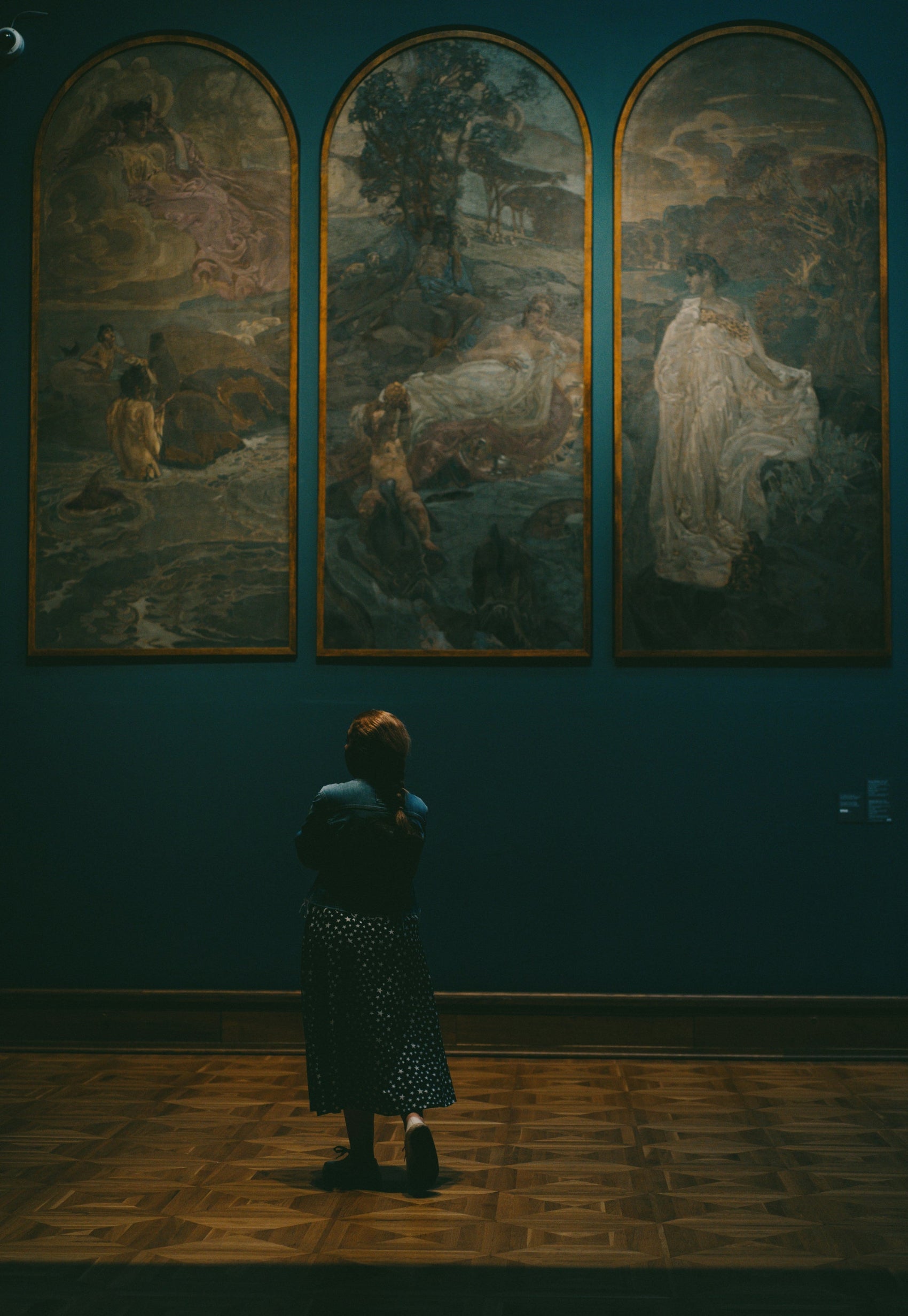 Person looking at three large paintings on a wall in a dimly lit room.